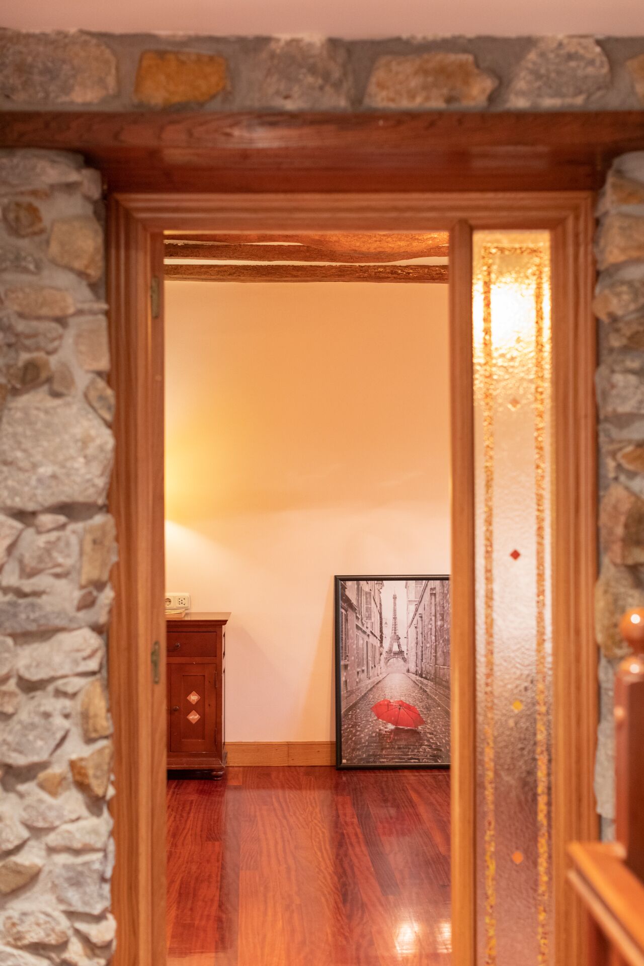 A wooden-framed doorway leading to a room with a stone wall, a wooden floor, and a framed picture on the wall.