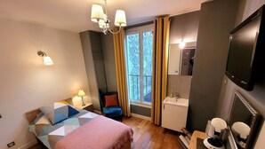 A hotel room with a bed, bedside table, lamp, chair, window with curtains, sink, mirror, and a television. - Hôtel oh sèvres autrement (Sevres)