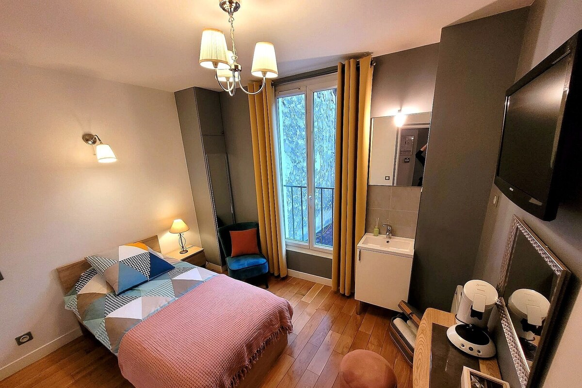 A hotel room with a bed, bedside table, lamp, chair, window with curtains, sink, mirror, and a television.