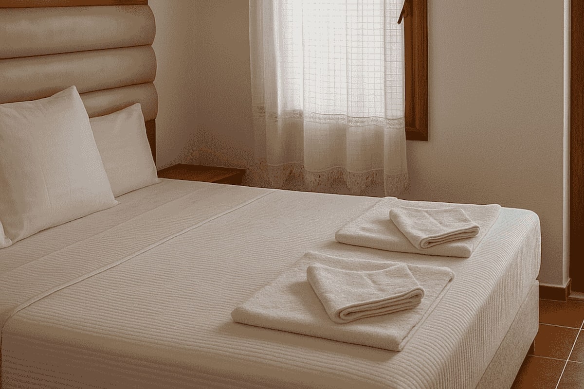 A neatly made bed with pillows and folded towels, a wooden headboard, and a window with sheer curtains.