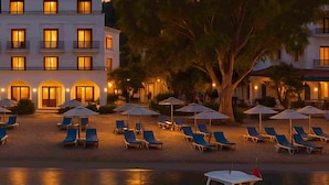 A beachfront hotel with sun loungers and umbrellas, a boat docked in the water, and a large tree in the foreground. - Antique İasos Butik Otel (Milas)
