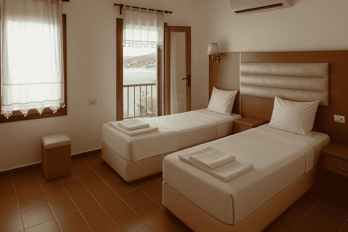 A hotel room with two single beds, a small bedside table, a nightstand, a window with curtains, and a view of the outdoors.