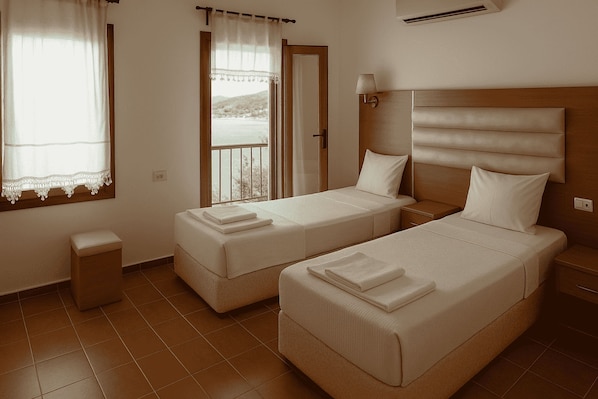 Habitación de hotel con dos camas individuales, una mesita de noche pequeña, cortinas en la ventana y vistas al exterior.