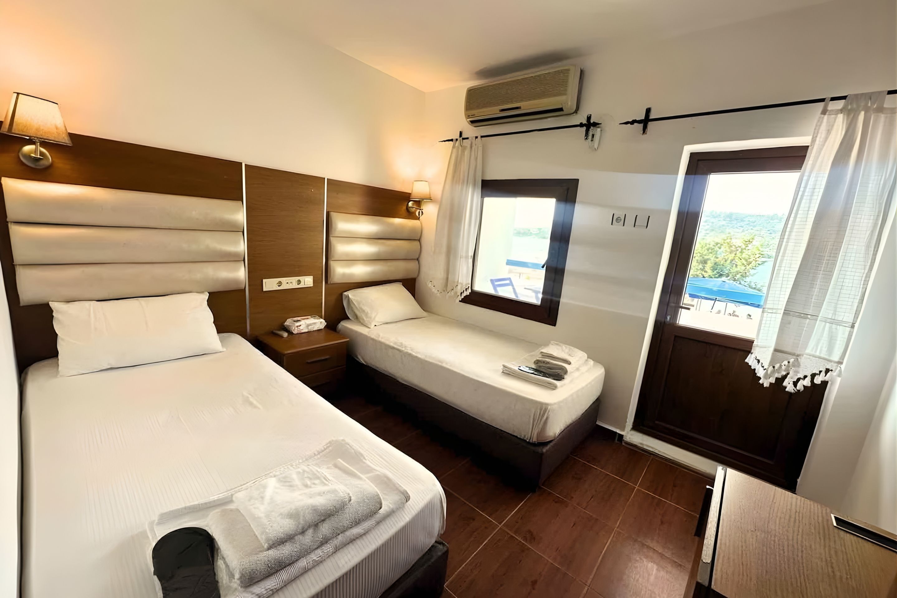 A hotel room with a large bed, a smaller bed, a bedside table with a lamp, a window with curtains, and an air conditioning unit.