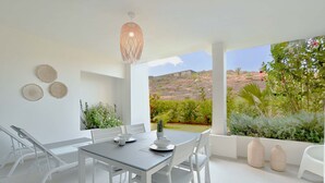 A modern dining area with a glass table, white chairs, and a view of a garden and hills.