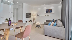 A modern living room with a grey sofa, a dining table with chairs, a TV mounted on the wall, and a ceiling-mounted air conditioner.