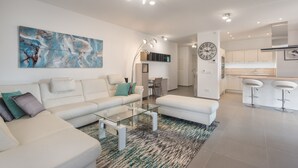 A modern living room with a white sofa, glass coffee table, and a large abstract painting. There is a kitchen area with white cabinets and a bar stool. - Colinas - Tinerfe 1.1 NATURE RESERVE VIEW 2B (Arona)