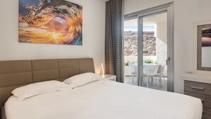 A bedroom with a large bed, a painting of a wave, a view of a desert landscape through a sliding door, and a small table with chairs. - Colinas - Tinerfe 1.1 NATURE RESERVE VIEW 2B (Arona)