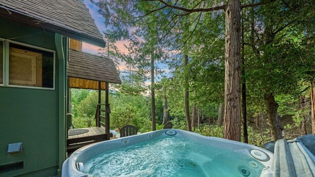 A hot tub in a forested area with a wooden deck and a house in the background. - Rocky Raccoon | Hot Tub, Lily Rock Views & Fireplace (Idyllwild)