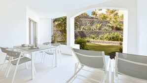 A modern dining area with white furniture, a stone wall, and a view of a garden. - Colinas - Tahona 0.2 POOL VIEW 1B (Arona)