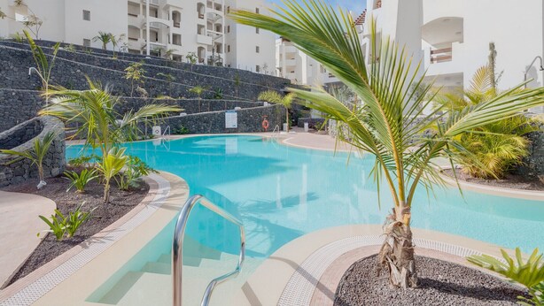 A swimming pool surrounded by palm trees and a stone wall, with white buildings in the background. - Colinas - Tahona 0.2 POOL VIEW 1B (Arona)