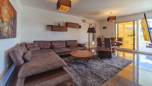 A modern living room with a sectional sofa, a round coffee table, and a large window offering a view of the outdoors. - Second Floor (Бјелиши)