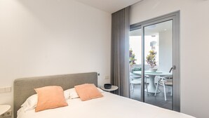 Une chambre d’hôtel avec un lit, une petite table sur laquelle se trouve un bol, et une vue sur un balcon extérieur agrémenté de chaises et de plantes.