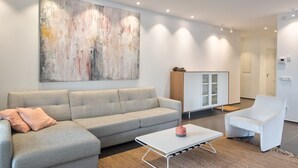 Un salon moderne avec un canapé gris, un fauteuil blanc et une table basse. Un grand tableau abstrait orne le mur, et on aperçoit un meuble avec des portes en verre.