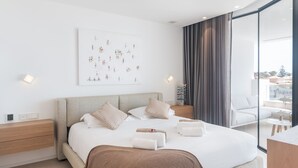 A modern hotel room with a large bed, a bedside table, a nightstand, a wall-mounted artwork, and a view of the outdoors through a sliding glass door. - Infinity 1.2 LUXE PARTIAL SEA VIEW 2B (Arona)