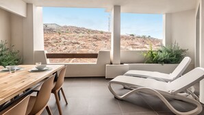 Een moderne kamer met een eettafel, stoelen, een witte fauteuil en uitzicht op een woestijnlandschap door grote ramen.