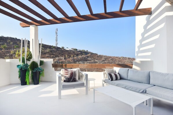 Un patio moderno al aire libre con un sofá blanco, una mesita y dos sillas. Hay plantas en macetas y una estructura de pérgola. El fondo muestra un paisaje montañoso con cielo despejado.