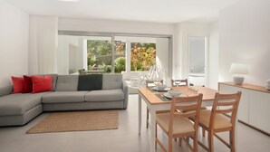 A modern living room with a grey sofa, red cushions, a wooden dining table, and a view of a garden through large windows. - Colinas -Bentor 0.3 POOL VIEW 1B  COLINAS (Arona)
