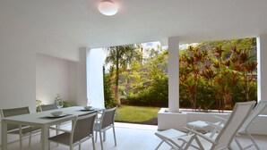A modern dining area with a white table and chairs, a glass of water, and a view of a lush garden through large windows. - Colinas -Bentor 0.3 POOL VIEW 1B  COLINAS (Arona)