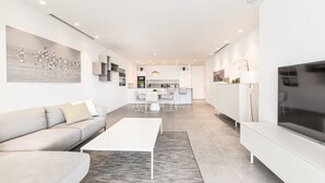 Un salon moderne avec un canapé, une table basse et un téléviseur. On aperçoit une cuisine à l’arrière-plan, avec un coin repas.
