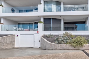 Un immeuble moderne à plusieurs étages, doté d’un garage, de balcons en verre et d’un espace paysager agrémenté de plantes.