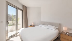Une chambre moderne avec un grand lit, une table de chevet, une lampe et une porte-fenêtre coulissante donnant sur un balcon.