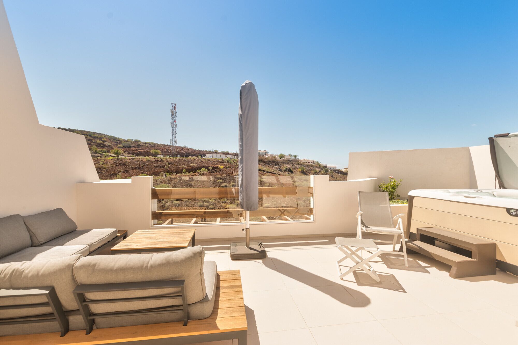 En moderne udendørs terrasse med sofa, spisebord og stole, med udsigt over et bakket landskab og et kommunikationstårn i det fjerne.