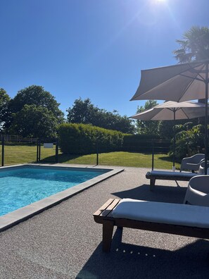 A swimming pool with lounge chairs and umbrellas, surrounded by a fence and greenery.