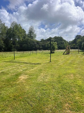 A grassy field with playground equipment, including swings and a slide, surrounded by trees and a clear sky.