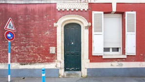 Un bâtiment en briques rouges, avec une porte verte et des volets blancs, un panneau de passage piéton et des bollards sur le trottoir.