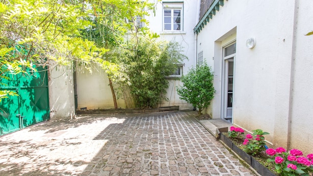 Une cour pavée de pierres, avec un bâtiment blanc, une clôture verte et des fleurs roses.
