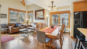 A spacious living room with a wooden dining table, a sofa, and a kitchen area with a refrigerator and cabinets. - Greyhawk Condos - Corner Unit - Private Hot Tub (Park City)