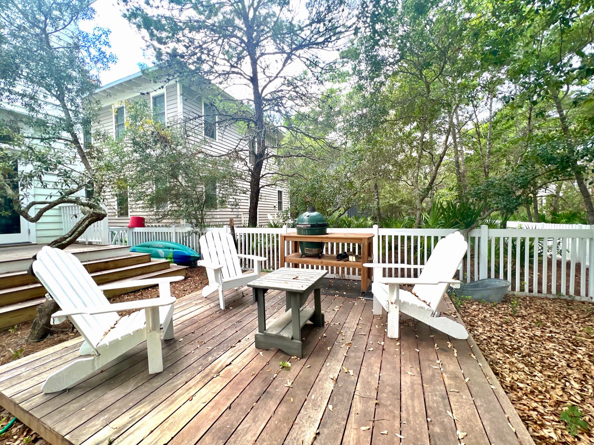 A wooden deck with white chairs and a table, surrounded by a white picket fence and trees.
