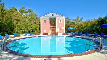 Una piscina con un edificio rojo al fondo, rodeada de sillas de descanso y sombrillas azules.