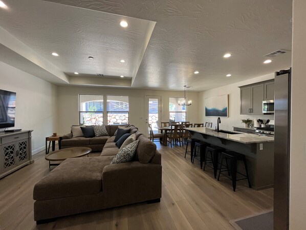 A spacious living room with a large sectional sofa, a dining area with a kitchen island, and a TV on a cabinet.