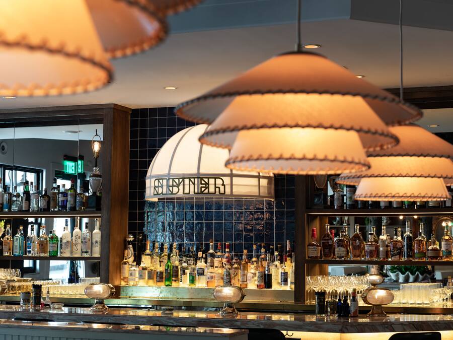 Zona de bar con varias estanterías llenas de botellas de licor, una barra bien iluminada y lámparas colgantes.