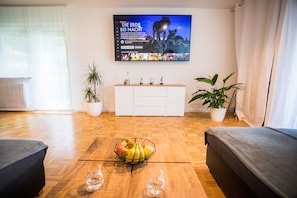 A living room with a TV displaying a movie, a wooden coffee table with fruit and glasses, a white cabinet, and two potted plants. - Auszeit im Schwarzwald (Lenzkirch)
