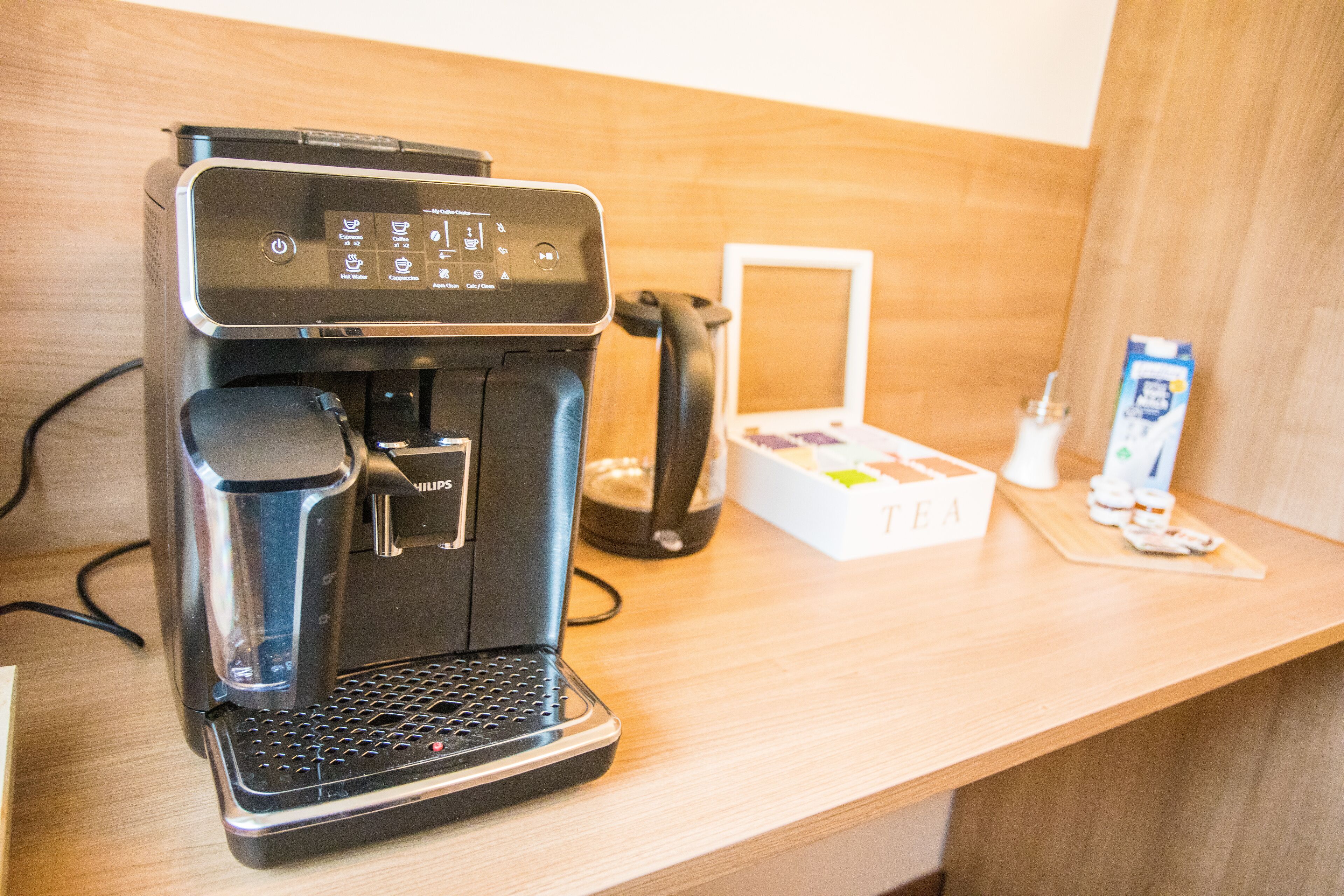 Une machine à café est posée sur un comptoir en bois, à côté d’un plateau avec du thé et d’une boîte de sachets de thé.