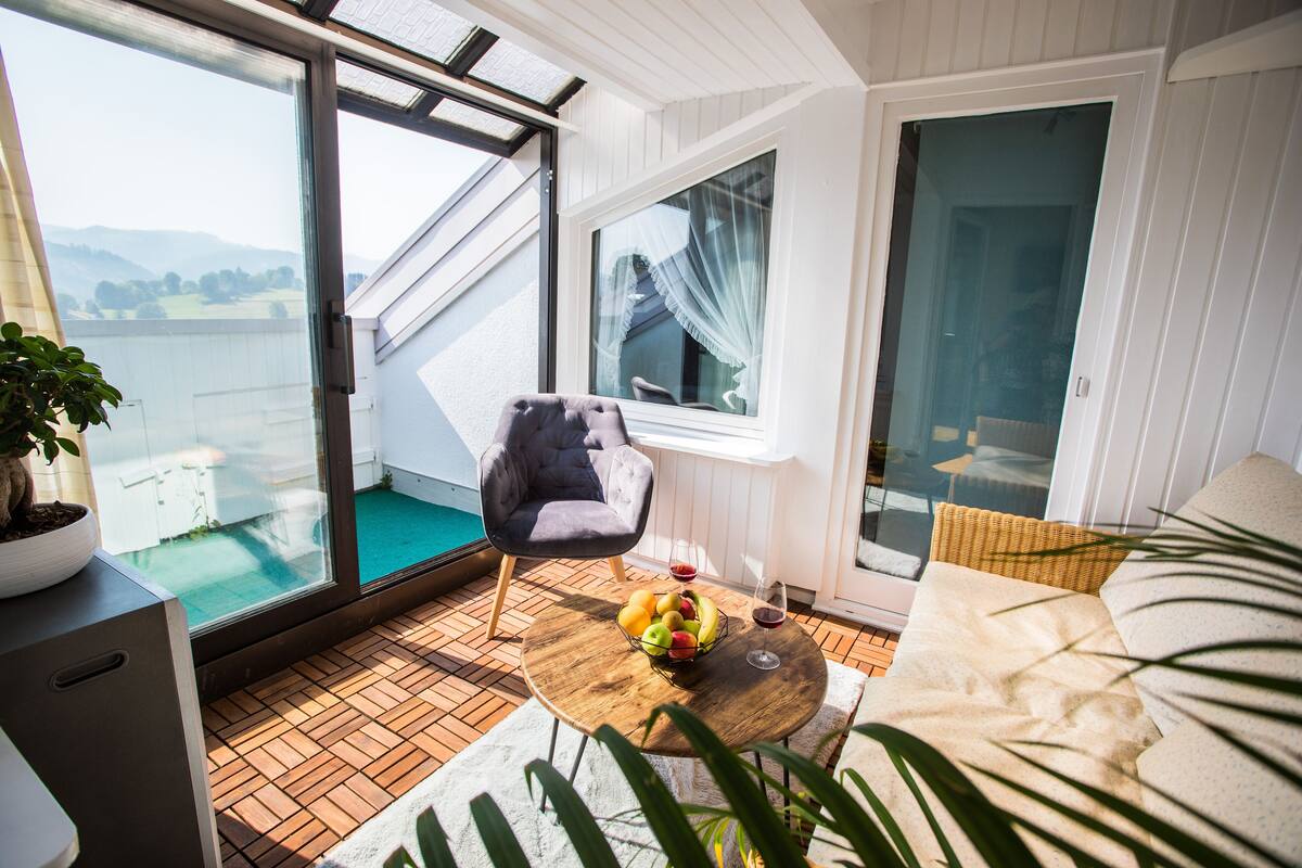 Un balcón con vistas al mar, una silla de mimbre, una mesa de madera con fruta y vino, y un sofá.