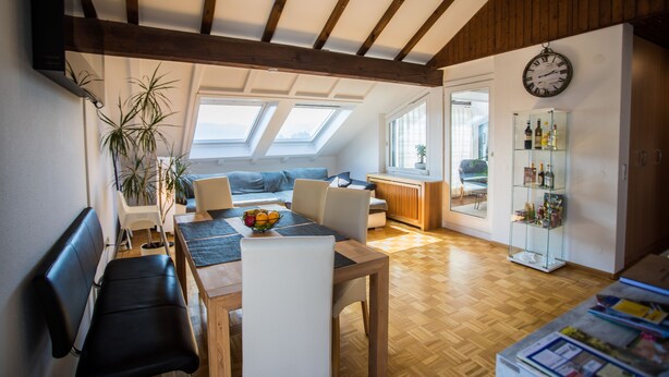 Ein modernes Wohnzimmer mit Holzboden, einem Essbereich mit Tisch und Stühlen, einer Couch und einem Bücherregal.