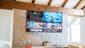 Ein Wohnzimmer mit einer Steinwand, einem Wand-Fernseher, der einen Film zeigt, einem kleinen Tisch mit einem Radio und einem Fenster.