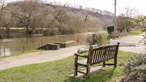 A bench by a river with a stone wall and a sign. - Steel Works Farm (Ross-on-Wye)