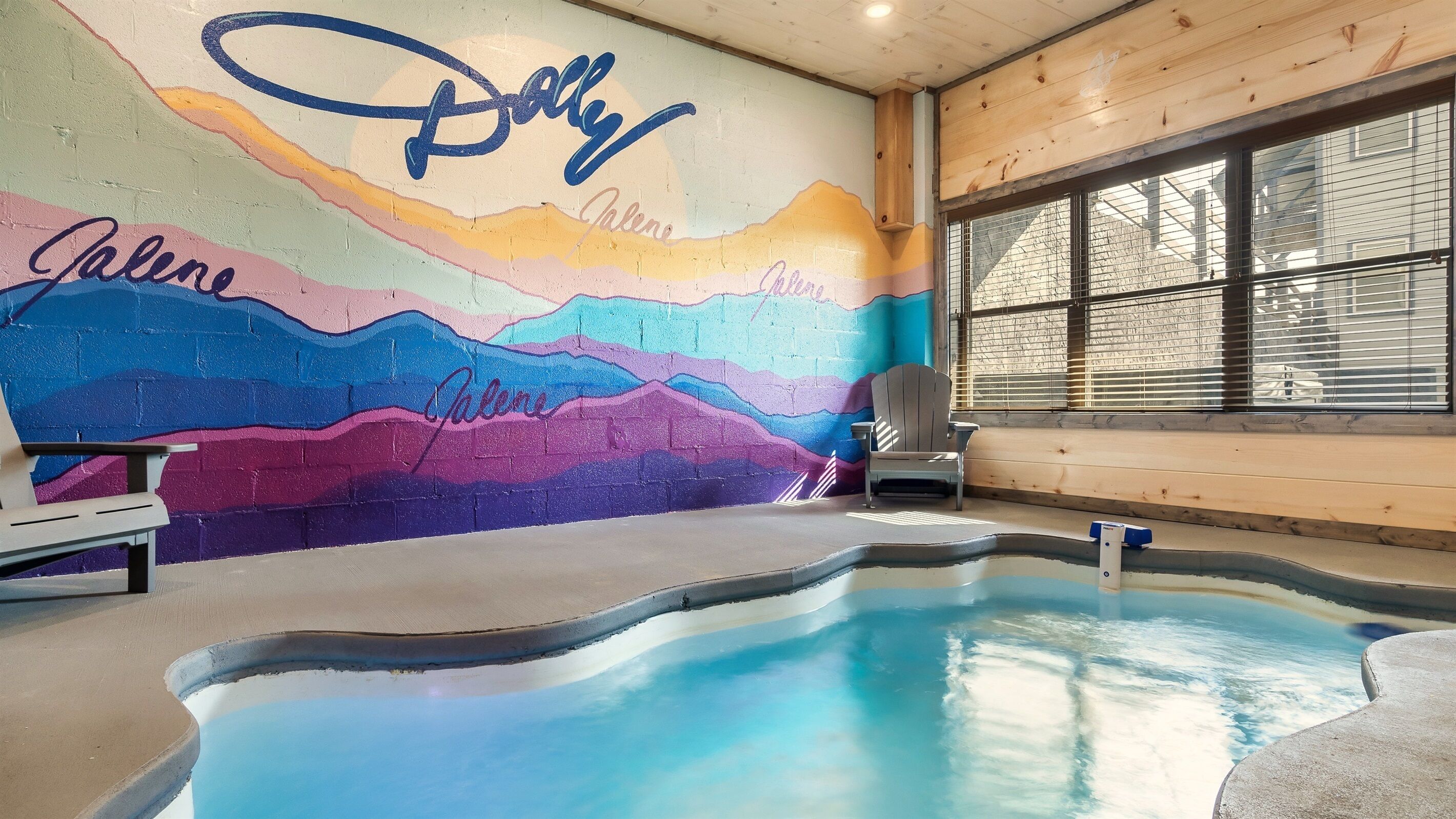 Ein Schwimmbecken mit Wandmalerei in bunten Farben, einem Stuhl und einem Fenster mit Jalousien.
