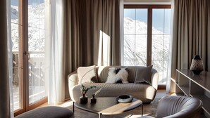 A modern living room with a sofa, coffee table, and armchair, large windows with curtains, and a view of snow-covered mountains.