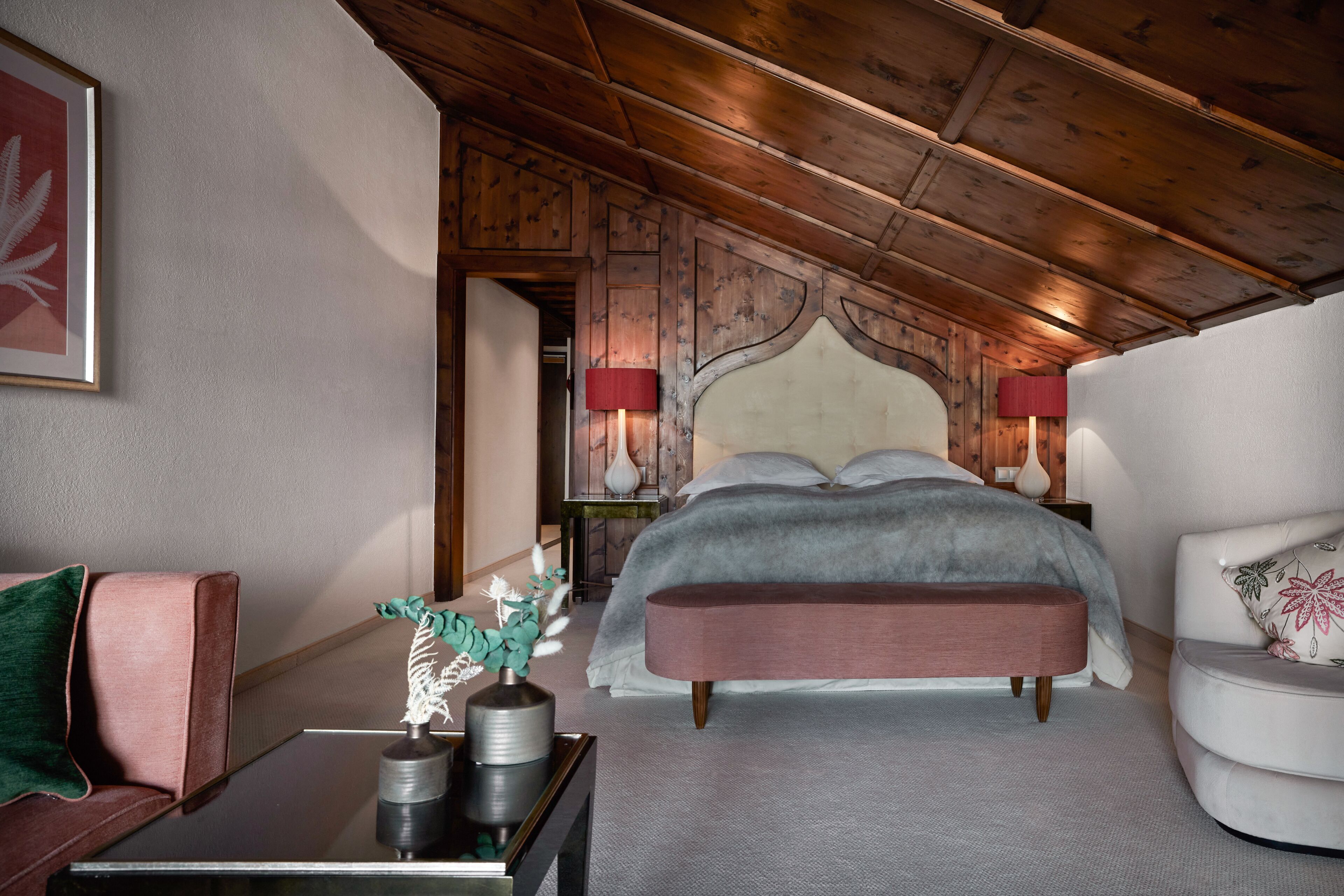 Une chambre avec un plafond en bois, un lit recouvert d’une couverture grise, un banc, une table de chevet avec des lampes et un tableau encadré au mur.