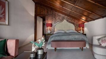 Une chambre avec un plafond en bois, un lit recouvert d’une couverture grise, un banc, une table de chevet avec des lampes et un tableau encadré au mur.