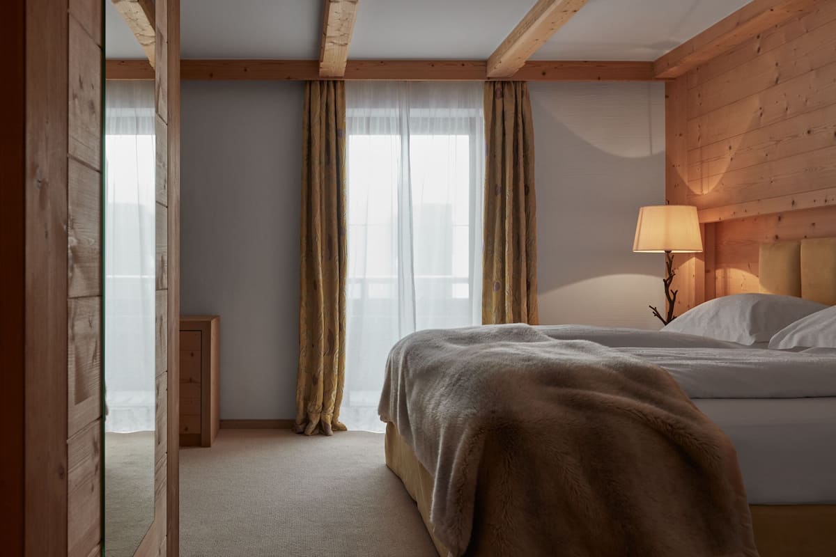 Un dormitorio con cama, cabecera de madera, mesita de noche con lámpara y cortinas traslúcidas.