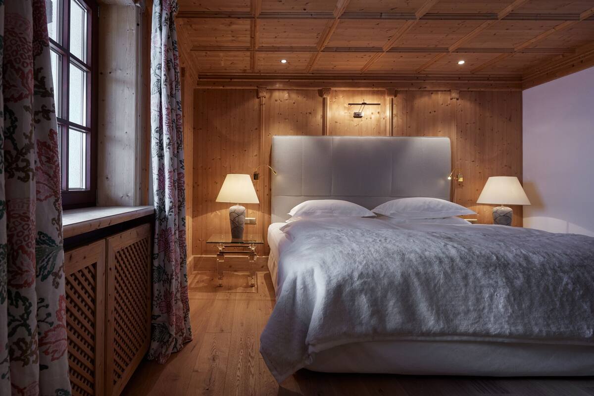 Un dormitorio con paneles de madera, una cama grande, dos lámparas de noche y un ventanal con cortinas de flores.