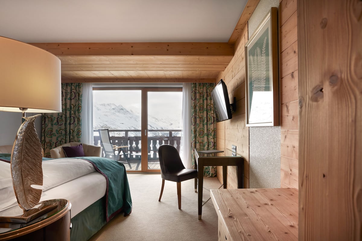 Habitación de hotel con cama, escritorio, silla, televisor y vistas a montañas nevadas a través de un ventanal grande.