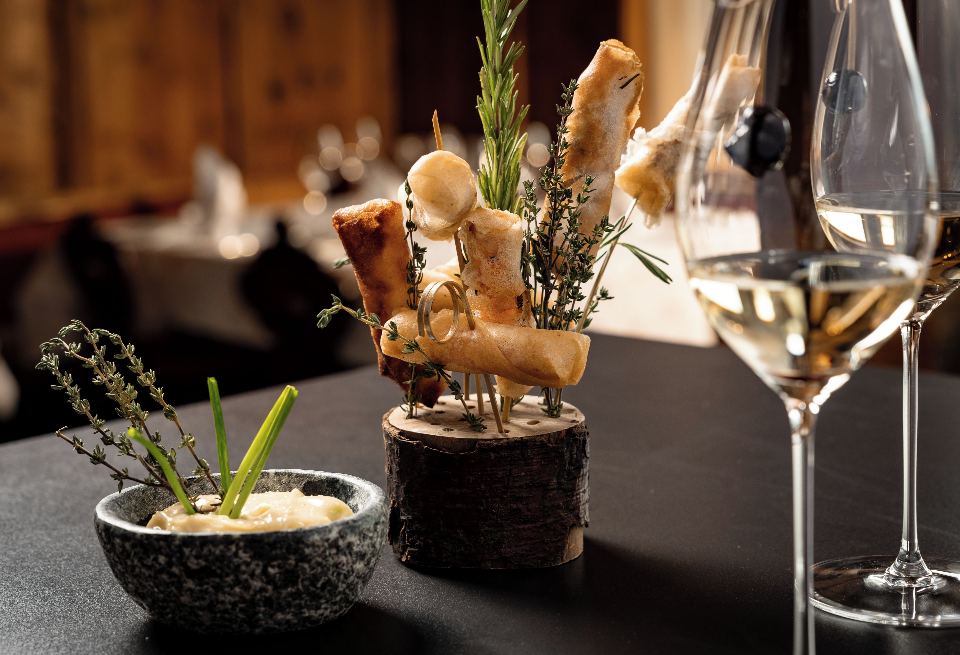 Una mesa dispuesta con una copa de vino blanco, un plato pequeño de comida y una decoración de palitos de pan y hierbas.